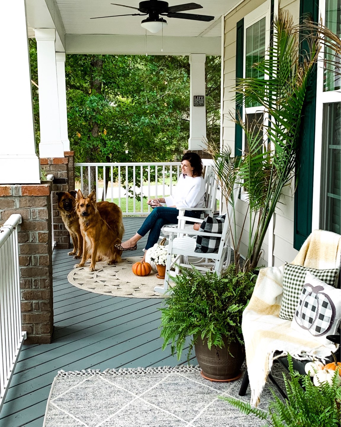 Our Inviting and Cozy Fall Front Porch Decor Ideas - I'm Fixin' To - @mbg0112 | Our Inviting and Cozy Fall Front Porch Decor Ideas by popular North Carolina lifestyle blog, I'm Fixin' To: image of a woman and her two dogs sitting on a front porch decorated with a Target Opalhouse Desert Hatch Outdoor Rug Gray, Amazon Jerimiah Spindleback BenchAmazon 4TH Emotion Set of 2 Farmhouse Buffalo Check Plaid Throw Pillow Covers, Target Smith and Hacken 12" Iron Galvanized Planters, Target Threshold 22" Artificial Berry Wreath White, pumpkins, Target Smart Living San Rafael II 15" LED Solar Mission Outdoor Lanterns, Hayneedle Coral Coast Indoor/Outdoor Mission Slat Rocking Chairs, mums, and green plants. 
