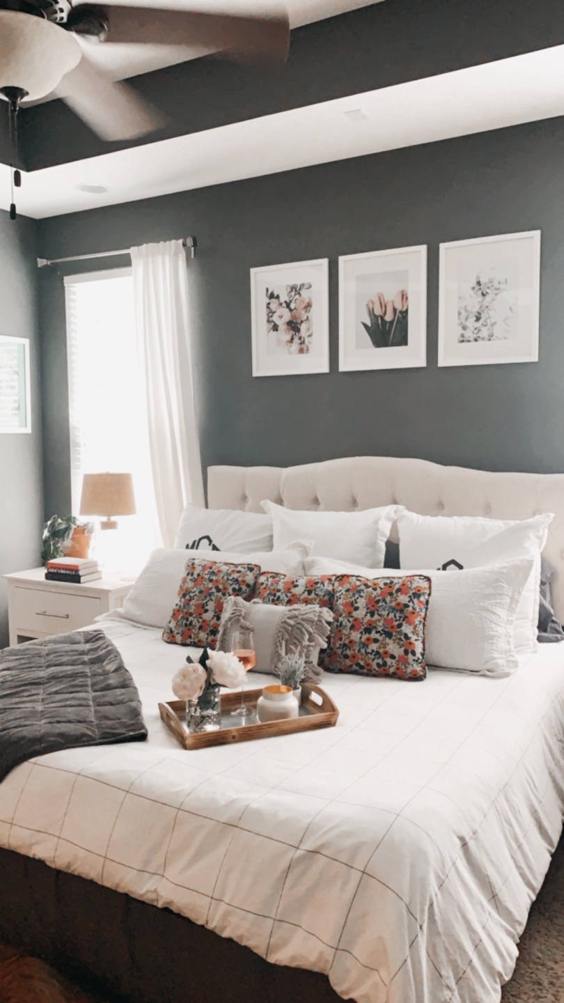 How to Make Your Home Cozy by popular NC life and style blog, I'm Fixin' To: image of a bedroom with a bed with a white tuft headboard ad white bedding, floral print artwork, white night stands, white curtains, grey walls, and a serving tray set with a vase of flowers and a candle. 