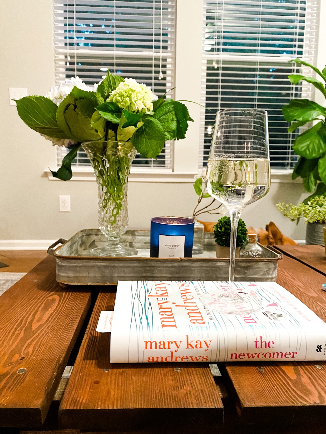 5 Affordable Ways to Keep your Home Feeling Fresh - I'm Fixin' To - @imfixintoblog | Ways to Refresh Your Home by popular NC life and style blog, I'm Fixin' To: image of a glass of white wine on top of "The Newcomer" book by Mary Kay Andrews next to a metal serving tray filled with a vase of white flowers and a blue candle votive. 
