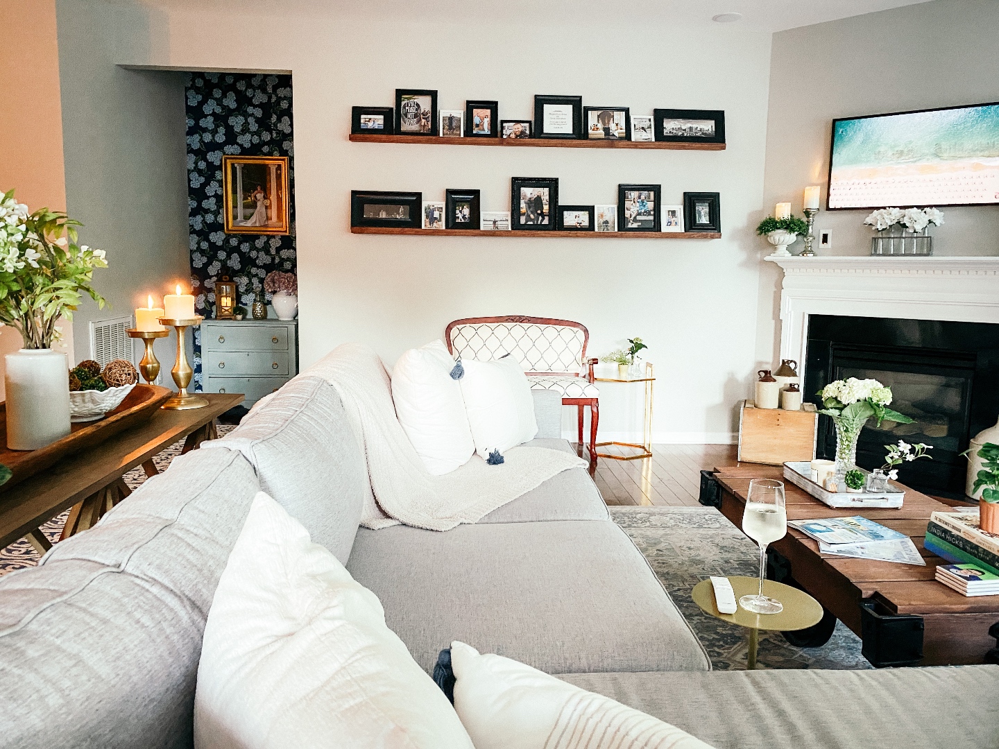 Room Reveal: Blue and Gray Living Room Ideas - I'm Fixin' To - @imfixintoblog | Blue and Grey Living Room Ideas by popular NC life and style blog, I'm Fixin' To: image of a living room decorated with a blue and white rug runner, light wood sofa table, house plants, floating wood shelves with black picture frames, white pillar candles, wooden picnic basket, blue and white floral wallpaper, and vintage blue and white armchair. 