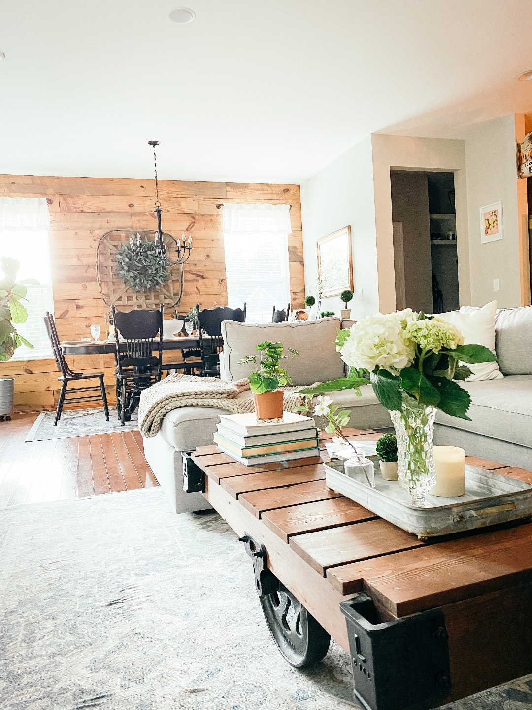 Room Reveal: Blue and Gray Living Room Ideas - I'm Fixin' To - @imfixintoblog | Blue and Grey Living Room Ideas by popular NC life and style blog, I'm Fixin' To: image of a living room decorated with a blue and white rug runner, light wood sofa table, house plants, floating wood shelves with black picture frames, white pillar candles, wooden picnic basket, blue and white floral wallpaper, cream cable knit throw blanket, and black dining table next to a shiplap accent wall. 