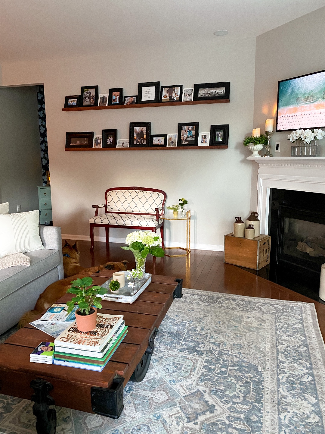 Room Reveal: Blue and Gray Living Room Ideas - I'm Fixin' To - @imfixintoblog | Blue and Grey Living Room Ideas by popular NC life and style blog, I'm Fixin' To: image of a living room decorated with a blue and white rug runner, light wood sofa table, house plants, floating wood shelves with black picture frames, white pillar candles, wooden picnic basket, blue and white floral wallpaper, and fireplace with white mantle. 