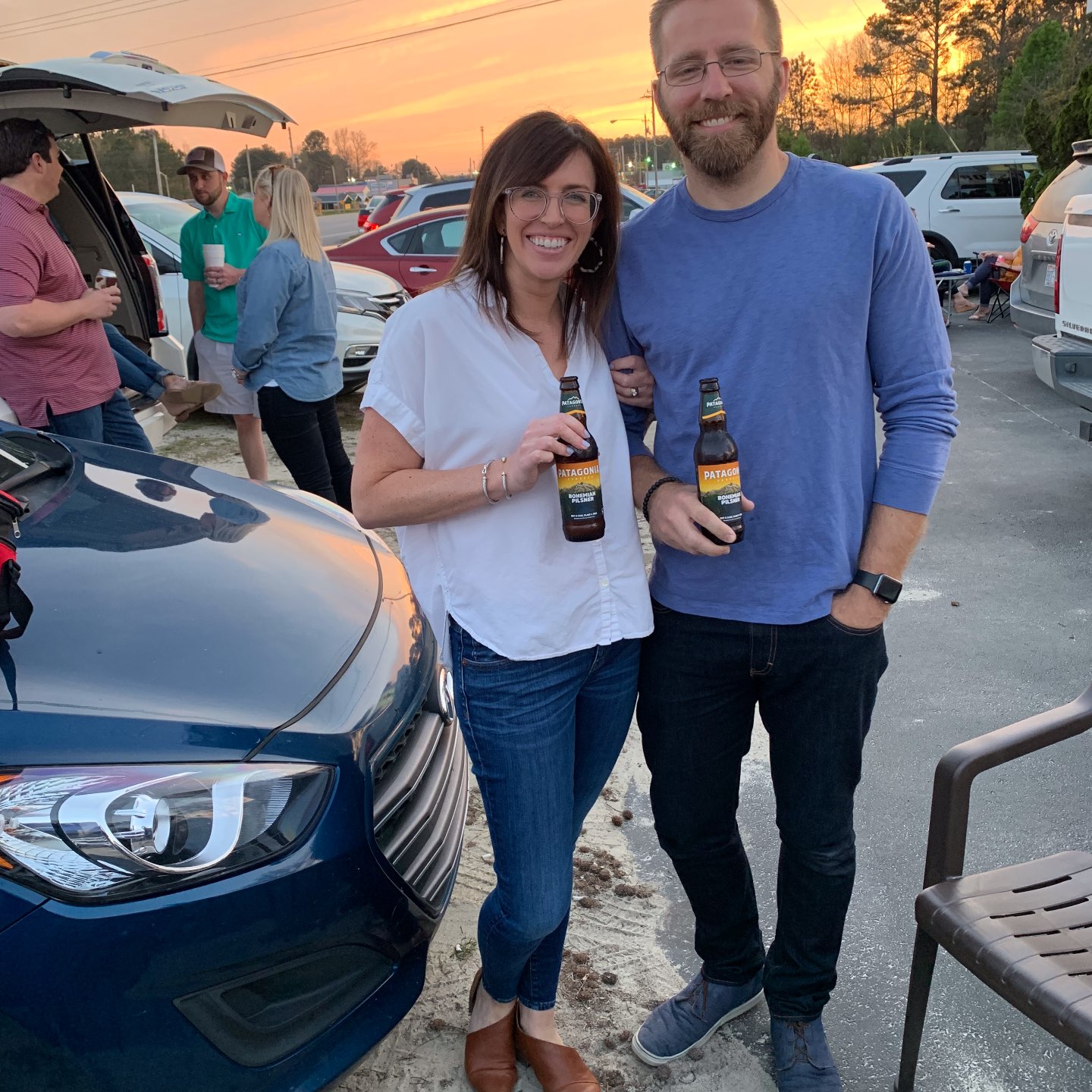 A couple stands happily in a parking lot at sunset, holding beer bottles labeled "Patagonia." They are smiling and dressed casually, surrounded by parked cars and friends.
